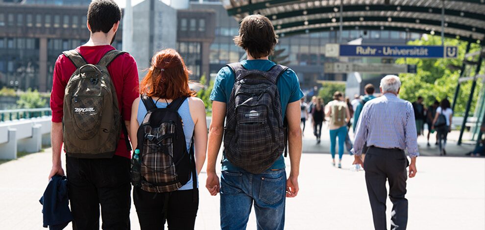 Drei Studierende sind von hinten abgebildet. Sie tragen sommerliche Kleidung und laufen auf der Unibrücke in Richtung der Bahnstation.